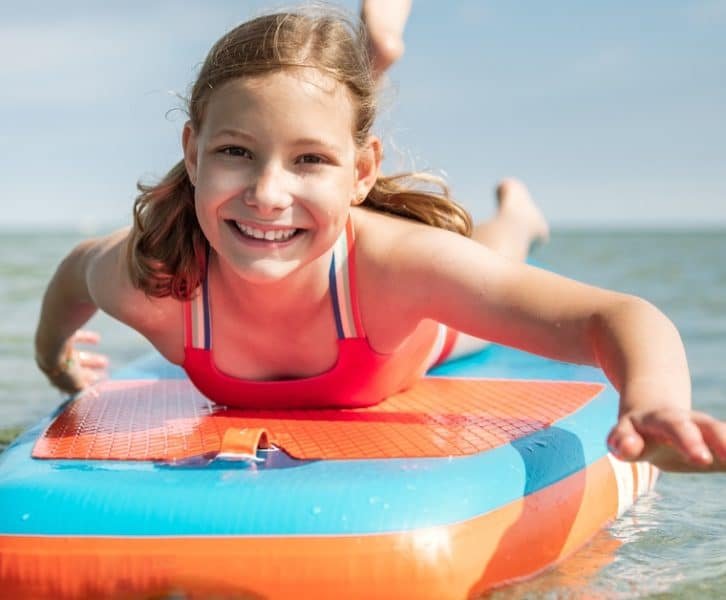 A young girl smiling toward the viewer as she rides on a blue-and-orange paddleboard on a large body of calm, open water.