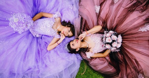 Two girls in poofy dresses, one purple and one pink, lie on the grass facing each other and making eye contact.