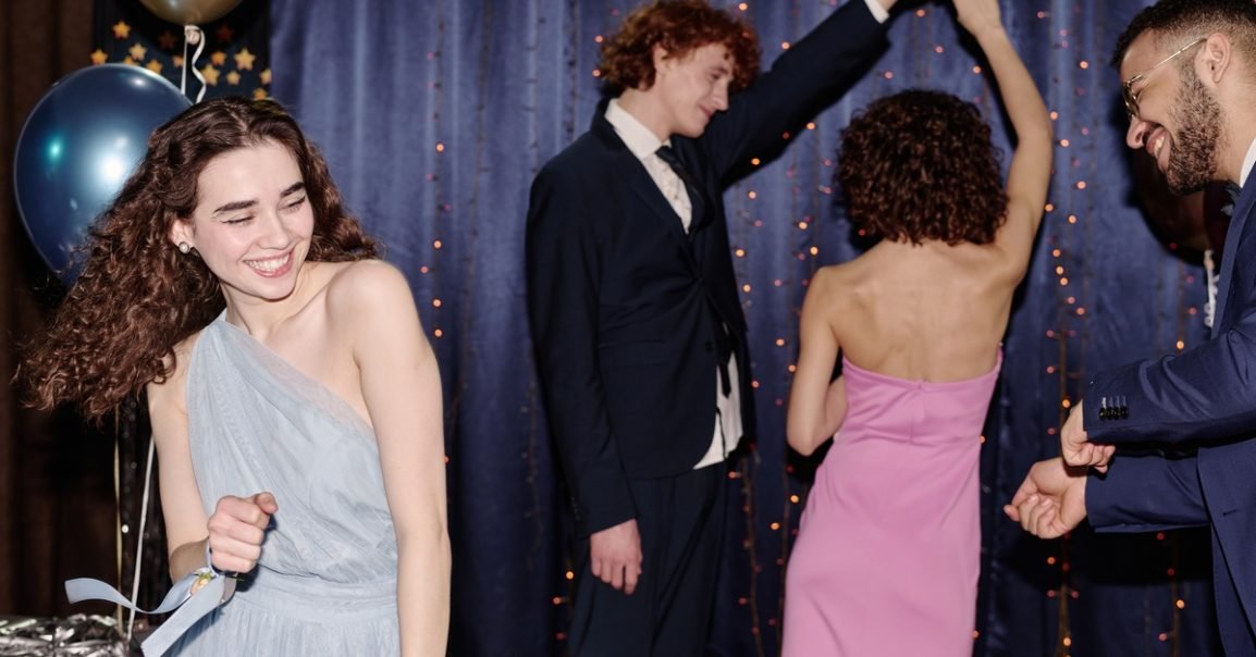 Four teenagers, including a girl in a light blue dress, dance and smile next to balloons and a blue drape backdrop with string lights.