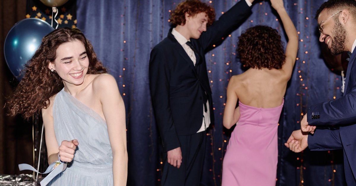 Four teenagers, including a girl in a light blue dress, dance and smile next to balloons and a blue drape backdrop with string lights.