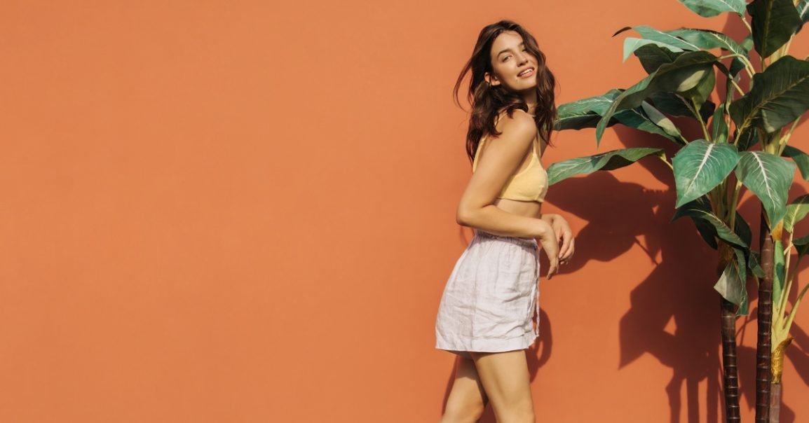 A teenage girl posing next to a tall plant against an orange wall. She wears white linen shorts and a yellow bralette-style top.